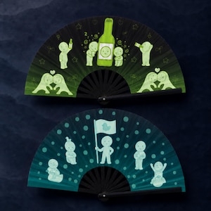 Puede incluir: Dos abanicos plegables con varillas negras. El abanico superior es verde oscuro con ilustraciones en verde claro de figuras, una botella y corazones. El abanico inferior es azul verdoso con ilustraciones en azul claro de figuras y una bandera.