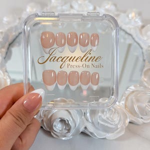 May include: A clear plastic case holds a set of press-on nails. The nails have a natural beige base with a white French tip. The text "Jacqueline Press-On Nails" is printed in gold script on the case.