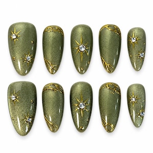 May include: A set of ten almond-shaped press-on nails in a shimmering olive green finish. The nails feature gold starburst accents with embedded clear rhinestones and raised gold metallic filigree patterns along the cuticle edges.