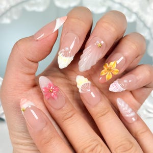May include: Almond-shaped artificial nails featuring a nude base with 3D embellishments including white seashell textures, small gold star studs, tiny white pearls, and hand-painted yellow and pink floral designs on select nails.