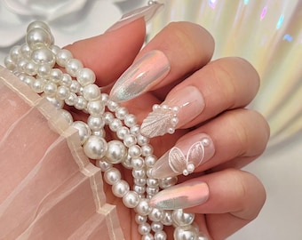 May include: Close-up of a hand with long, almond-shaped nails. The nails are painted with a pearlescent white polish and adorned with delicate white 3D floral designs and pearl accents.