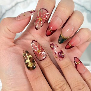 May include: A hand displaying a set of long, almond-shaped nails with intricate nail art. The designs include floral patterns, gold accents, and a gradient of colors, including red, pink, and brown. Some nails feature 3D floral embellishments.