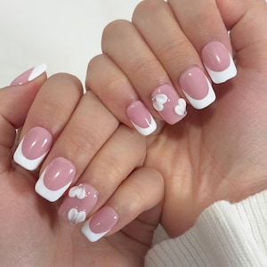 May include: A set of manicured nails featuring a pink base with white tips in a French manicure style. Some nails have 3D white heart-shaped embellishments and small silver accents. The nails are on a light skin tone.