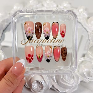 May include: A set of press-on nails in a clear plastic case. The nails feature various designs, including floral accents, gold studs, and a tortoiseshell pattern. The text "Jacqueline Press On Nails" is visible on the case.