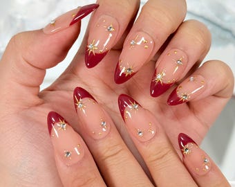 May include: A hand displaying long, almond-shaped nails. The nails feature a clear base with a deep red French tip. Gold starburst and small crystal accents are applied to the nails.