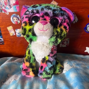 Könnte beinhalten: Ein Plüsch-Leopard mit einem leuchtend bunten Fellmuster in Neon-Grün, Pink, Blau und Gelb mit schwarzen Flecken. Das Stofftier hat große, glitzernde dunkle Augen, eine kleine rosa Nase und einen weißen Fleck auf der Brust.