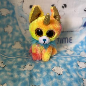 May include: A colourful plush toy, a hybrid of a unicorn and a dog. It features large, glittery eyes, a gold horn, and gold glitter ears. The fur is a blend of yellow, orange, and pink. It has a blue collar and a blue nose.