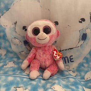 May include: A pink plush monkey toy with large, black eyes and a white face. The monkey has a red heart-shaped tag attached. It is sitting on a blue and white patterned fabric.