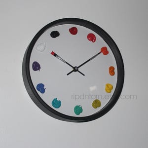 May include: A round wall clock with a black frame and a white face. Instead of numbers, the clock features twelve distinct dollops of colorful paint arranged in a circle. The hour and minute hands are shaped like thin paintbrushes.