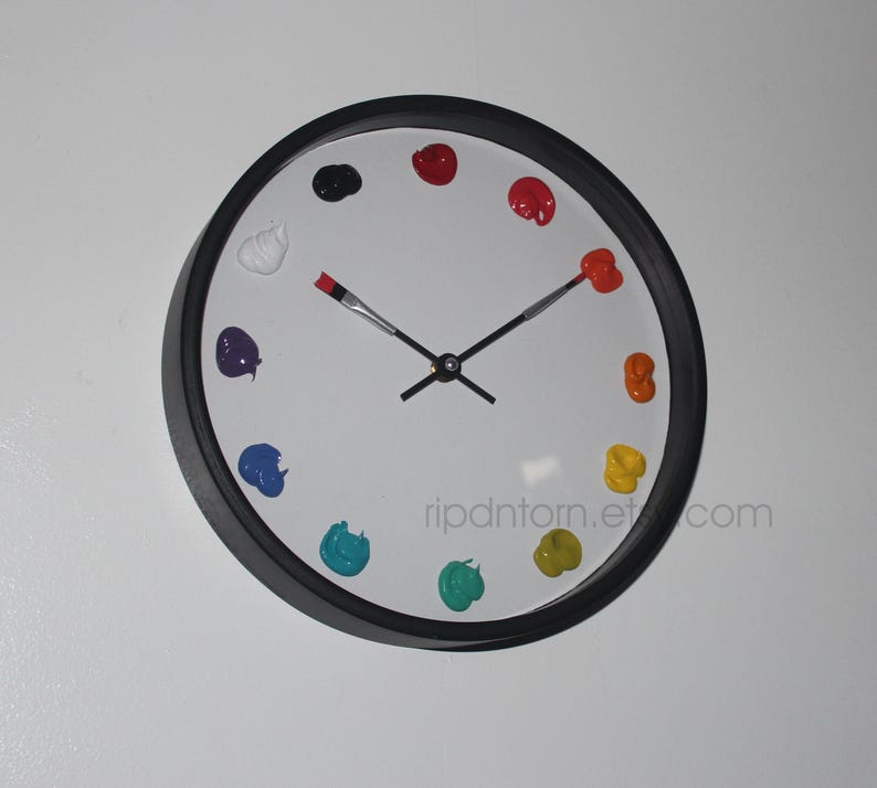 May include: A round wall clock with a black frame and a white face. Instead of numbers, the clock features twelve distinct dollops of colorful paint arranged in a circle. The hour and minute hands are shaped like thin paintbrushes.