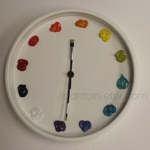May include: A round white wall clock featuring twelve distinct dollops of colorful paint serving as hour markers. The clock hands are represented by a single paintbrush with a black handle and green bristles. The text ripdntorn.etsy.com is visible.