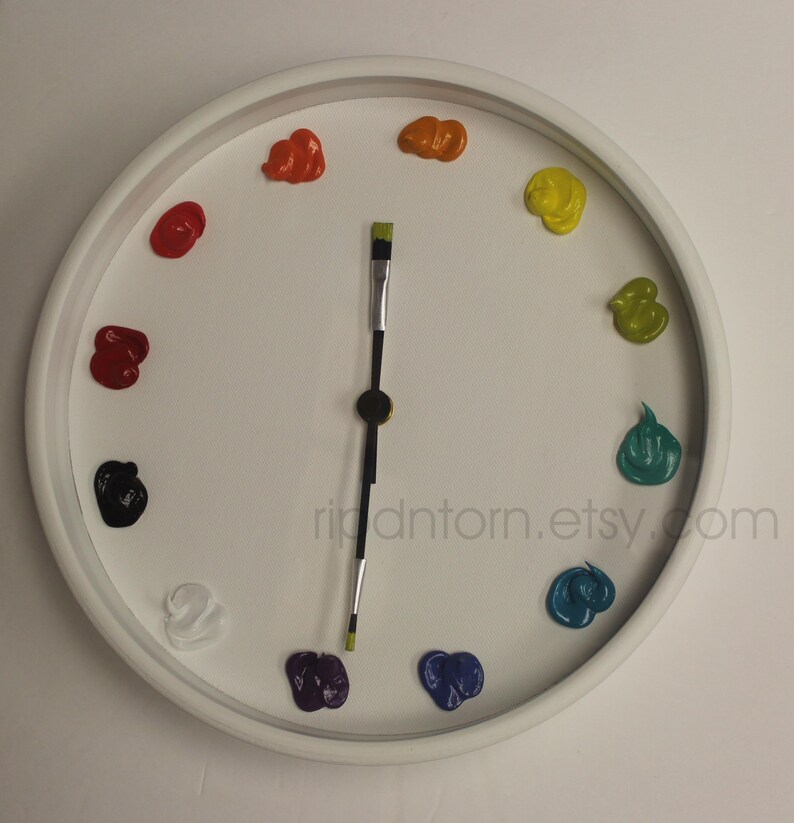 May include: A round white wall clock featuring twelve distinct dollops of colorful paint serving as hour markers. The clock hands are represented by a single paintbrush with a black handle and green bristles. The text ripdntorn.etsy.com is visible.