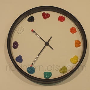 May include: A round wall clock with a black frame and a white face. Instead of numbers, the clock features twelve distinct dollops of colorful paint arranged in a circle. The hour and minute hands are shaped like small paintbrushes.