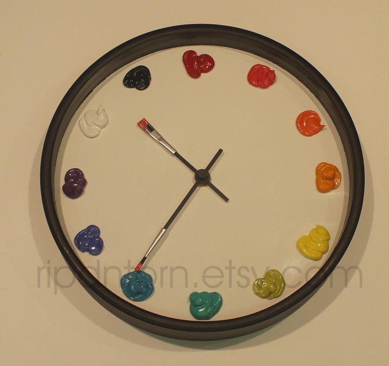 May include: A round wall clock with a black frame and a white face. Instead of numbers, the clock features twelve distinct dollops of colorful paint arranged in a circle. The hour and minute hands are shaped like small paintbrushes.