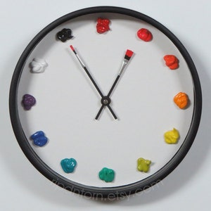 May include: A round wall clock with a black frame and a white face. Instead of numbers, twelve distinct dollops of colorful paint mark the hours. The clock hands are shaped like two thin paintbrushes, one with a red tip, pointing to the time.