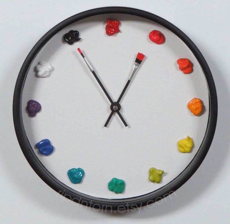 May include: A round wall clock with a black frame and a white face. Instead of numbers, twelve distinct dollops of colorful paint mark the hours. The clock hands are shaped like two thin paintbrushes, one with a red tip, pointing to the time.