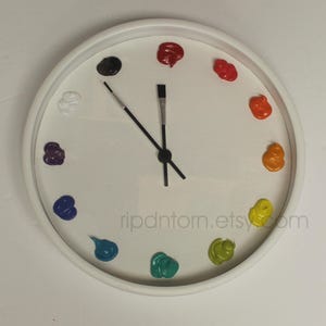 May include: A white wall clock with a circular frame and a white face. Instead of numbers, twelve distinct dollops of colorful paint are arranged in a circle. The hour and minute hands are shaped like thin black paintbrushes with silver ferrules.