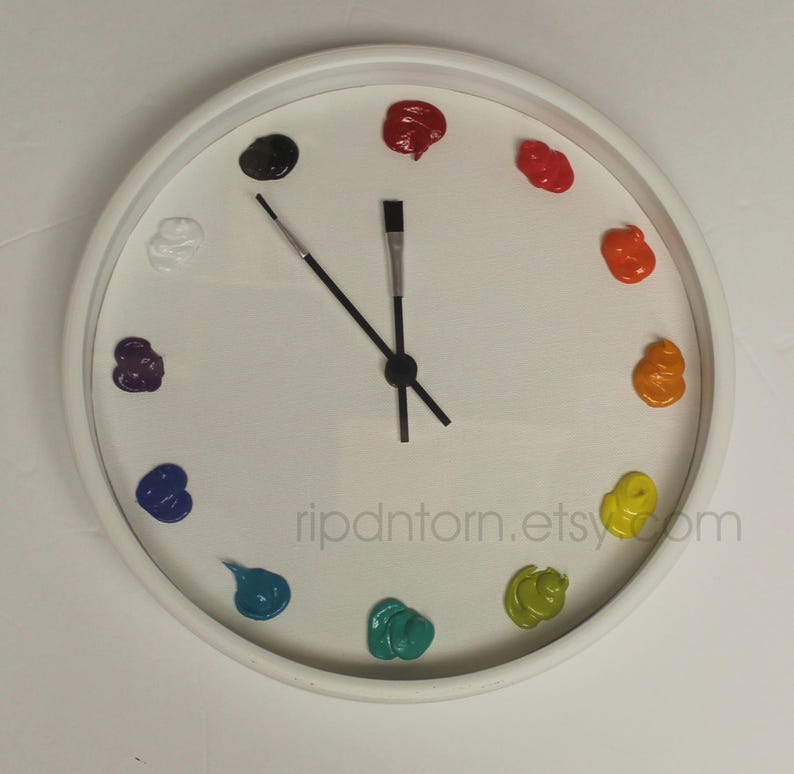 May include: A white wall clock with a circular frame and a white face. Instead of numbers, twelve distinct dollops of colorful paint are arranged in a circle. The hour and minute hands are shaped like thin black paintbrushes with silver ferrules.