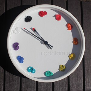 May include: A round white wall clock with a white face featuring twelve distinct dollops of colorful paint arranged as hour markers. The clock hands are shaped like two thin paintbrushes with black bristles and red tips, pointing to the ten and two.