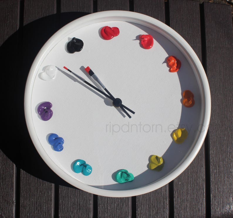 May include: A round white wall clock with a white face featuring twelve distinct dollops of colorful paint arranged as hour markers. The clock hands are shaped like two thin paintbrushes with black bristles and red tips, pointing to the ten and two.