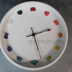 May include: A round white wall clock featuring twelve dollops of colorful oil paint as hour markers. The clock hands are shaped like thin paintbrushes with black handles and metal ferrules. The text ripntorn.etsy.com is printed on the face.