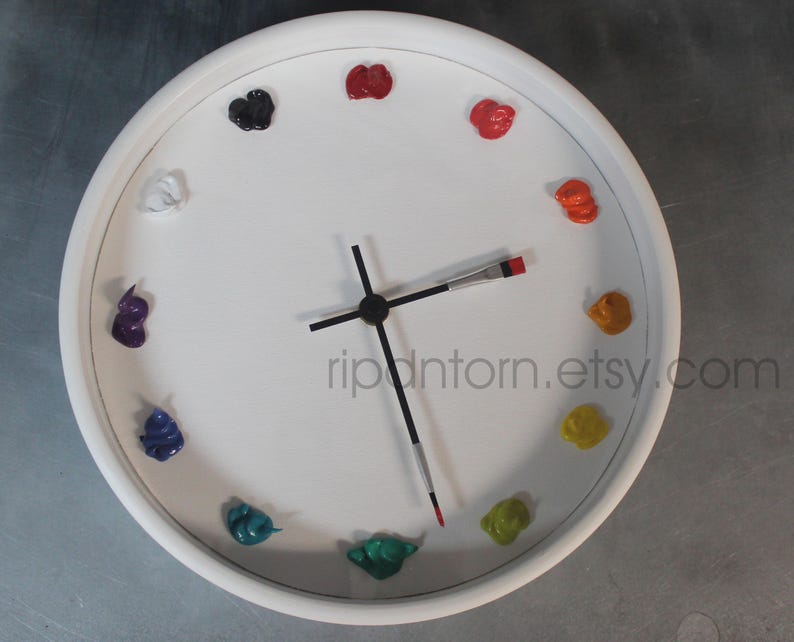 May include: A round white wall clock featuring twelve dollops of colorful oil paint as hour markers. The clock hands are shaped like thin paintbrushes with black handles and metal ferrules. The text ripntorn.etsy.com is printed on the face.