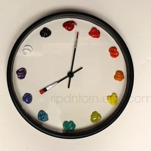 May include: A round wall clock with a black frame and a white face. Instead of numbers, twelve distinct dollops of colorful paint mark the hours. The clock hands are shaped like thin paintbrushes with black handles and silver ferrules.