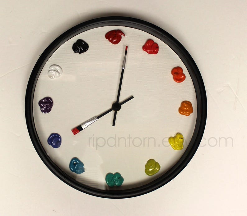 May include: A round wall clock with a black frame and a white face. Instead of numbers, twelve distinct dollops of colorful paint mark the hours. The clock hands are shaped like thin paintbrushes with black handles and silver ferrules.