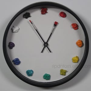 May include: A round wall clock with a black frame and a white face. Instead of numbers, the clock features twelve distinct dollops of colorful paint arranged in a circle. The hour and minute hands are shaped like thin paintbrushes.