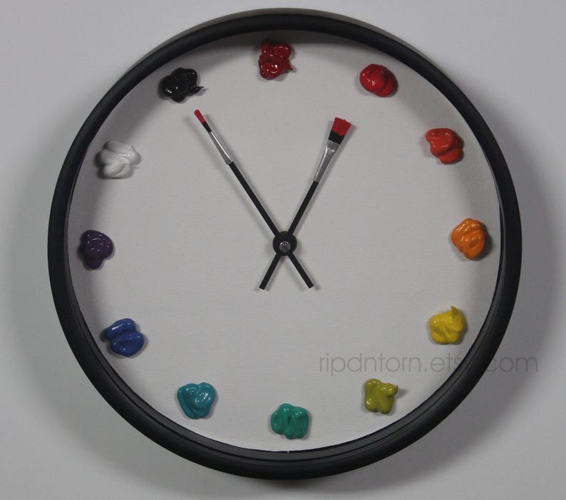 May include: A round wall clock with a black frame and a white face. Instead of numbers, the clock features twelve distinct dollops of colorful paint arranged in a circle. The hour and minute hands are shaped like thin paintbrushes.