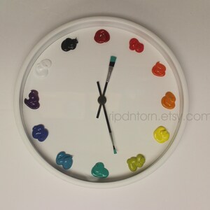 May include: A round white wall clock featuring twelve dollops of colorful paint as hour markers, including black, red, orange, yellow, green, teal, blue, purple, and white. The clock hands are represented by two thin paintbrushes.
