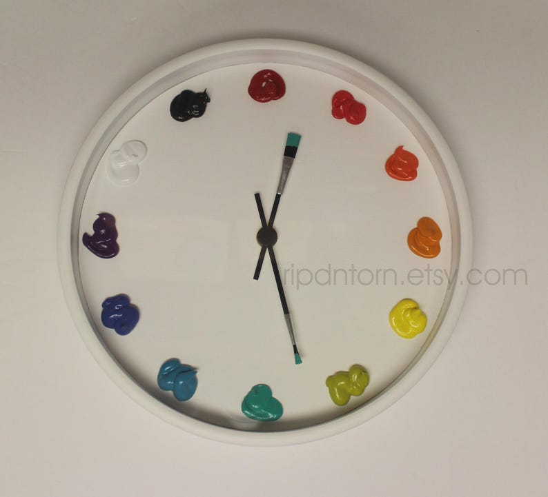 May include: A round white wall clock featuring twelve dollops of colorful paint as hour markers, including black, red, orange, yellow, green, teal, blue, purple, and white. The clock hands are represented by two thin paintbrushes.