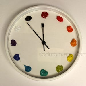 May include: A round white wall clock with a white face featuring twelve dollops of colorful paint serving as hour markers. The clock hands are shaped like two thin black paintbrushes. The text ripdntorn.etsy.com is printed in light gray.