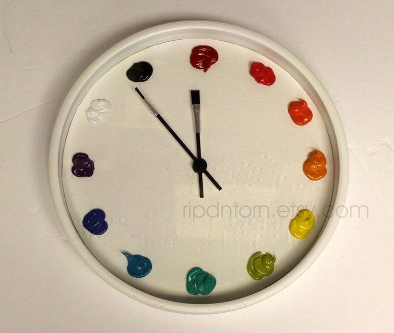May include: A round white wall clock with a white face featuring twelve dollops of colorful paint serving as hour markers. The clock hands are shaped like two thin black paintbrushes. The text ripdntorn.etsy.com is printed in light gray.