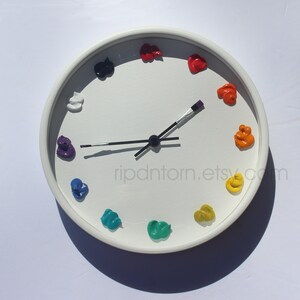 May include: A round white wall clock featuring twelve dollops of colorful paint as hour markers. The clock hands are shaped like thin paintbrushes with black handles and silver ferrules. The text ripdntorn.etsy.com is printed on the clock face.