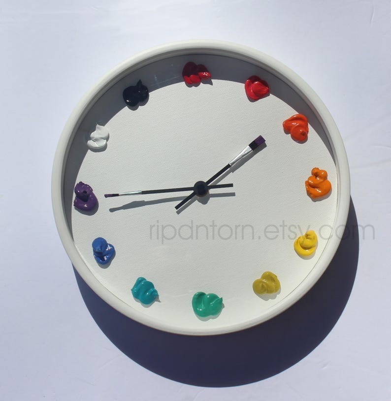 May include: A round white wall clock featuring twelve dollops of colorful paint as hour markers. The clock hands are shaped like thin paintbrushes with black handles and silver ferrules. The text ripdntorn.etsy.com is printed on the clock face.
