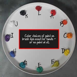 May include: A circular white palette featuring twelve distinct dollops of colored paint arranged in a clock-like pattern, each labeled with a letter from A to L. A central black rectangular sign displays the text: Color choices of paint on brush tips used for hands &ndash; or no paint at all.