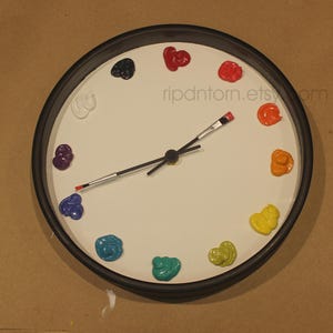 May include: A round wall clock with a black frame and a white face. Instead of numbers, twelve dollops of colorful paint mark the hours. The clock hands are shaped like thin paintbrushes with black handles and red tips. The text ripdntorn.etsy.com is visible.