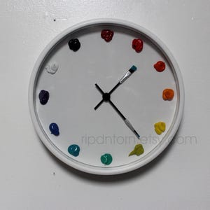 May include: A round white wall clock featuring twelve dollops of colorful paint as hour markers. The clock hands are shaped like two paintbrushes with black bristles and silver ferrules. The text ripdntorn.etsy.com is printed on the clock face.