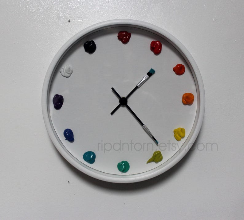May include: A round white wall clock featuring twelve dollops of colorful paint as hour markers. The clock hands are shaped like two paintbrushes with black bristles and silver ferrules. The text ripdntorn.etsy.com is printed on the clock face.