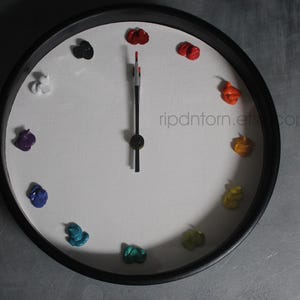 May include: A round wall clock with a black frame and a white face. Instead of numbers, twelve distinct dollops of colorful oil paint mark the hours. The clock features black hour and minute hands, with a red tip on the minute hand. Text reads ripdntorn.etsy.com.