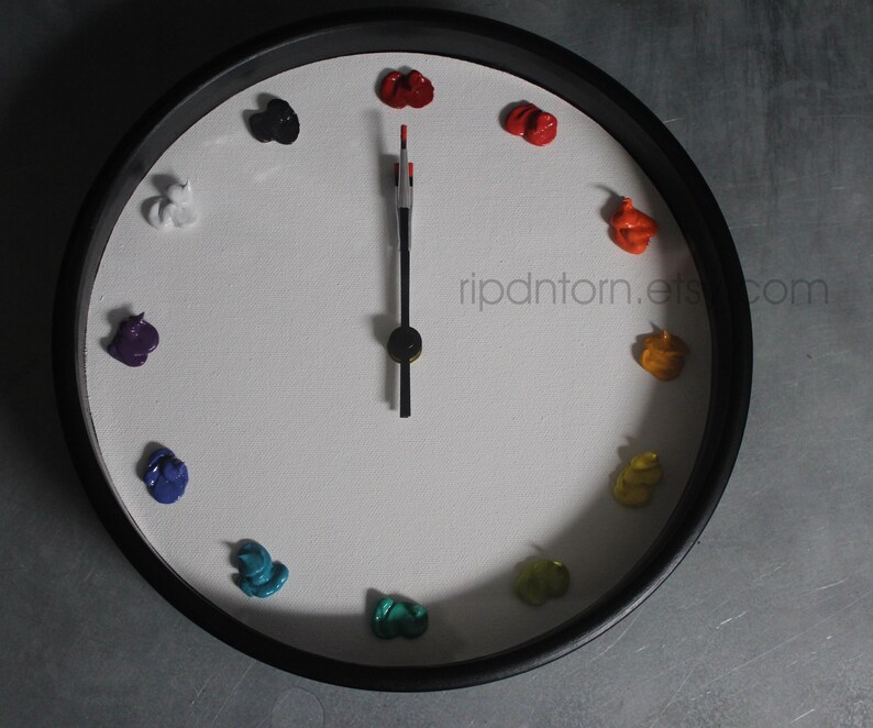 May include: A round wall clock with a black frame and a white face. Instead of numbers, twelve distinct dollops of colorful oil paint mark the hours. The clock features black hour and minute hands, with a red tip on the minute hand. Text reads ripdntorn.etsy.com.