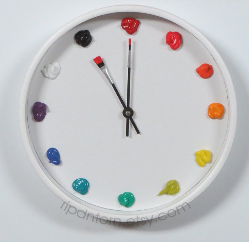 May include: A round white wall clock with a white face featuring twelve dollops of colorful paint as hour markers. The hour hand is a paintbrush with a red tip, and the minute hand is a thin black rod. The text ripdantorn.etsy.com is printed at the bottom.