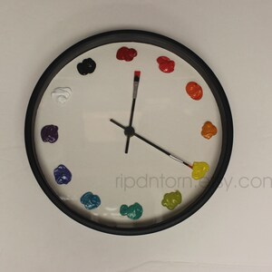 May include: A round wall clock with a black frame and a white face. Instead of numbers, twelve dollops of colorful paint mark the hours. The hour and minute hands are shaped like thin paintbrushes with black handles and metal ferrules.