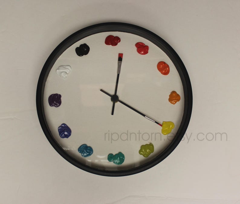 May include: A round wall clock with a black frame and a white face. Instead of numbers, twelve dollops of colorful paint mark the hours. The hour and minute hands are shaped like thin paintbrushes with black handles and metal ferrules.