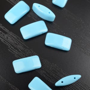 Könnte beinhalten: Eine Sammlung hellblauer rechteckiger und ovaler Perlen. Die Perlen sind glatt und glänzend, wobei zwei ovale Perlen Löcher zum Auffädeln haben. Die Perlen sind auf einer dunklen, strukturierten Oberfläche verstreut.