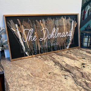 Può includere: Un'insegna incorniciata in legno con la scritta "The Dohlmans EST. 2022" in corsivo bianco. L'insegna presenta erbe e fiori secchi nei toni del marrone, beige e verde su sfondo nero.