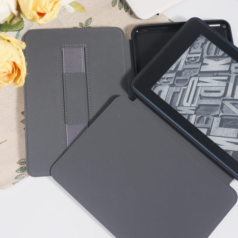 May include: A grey e-reader case with a hand strap and a separate cover. The e-reader screen displays a grey and white pattern. The case is made of a smooth material.