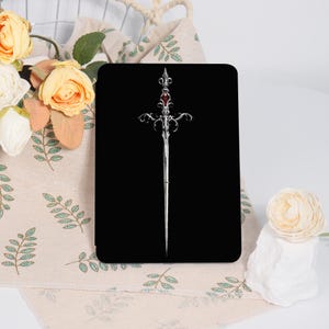 May include: A black rectangular product with a detailed silver sword design. The sword features an ornate hilt with a red gem and a long, slender blade. The design is centered on the product.