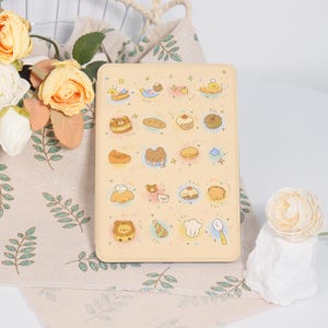 May include: A rectangular, light beige e-reader case featuring a repeating pattern of small, colorful illustrations including bears, various baked goods, stars, and kitchen utensils arranged in a grid across the front surface.